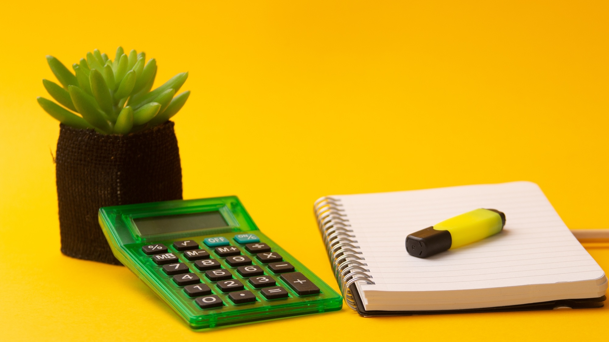 Selective focus shot of highlighter on a journal, calculator, and a plant on a yellow background