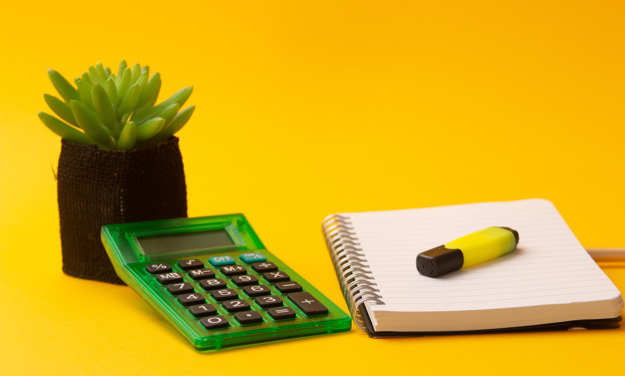 Selective focus shot of highlighter on a journal, calculator, and a plant on a yellow background