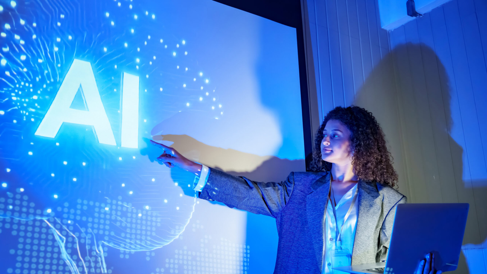A woman is explaining artificial intelligence concepts in a contemporary office. She uses a laptop and points to a bright screen displaying blue visual elements related to AI technology.