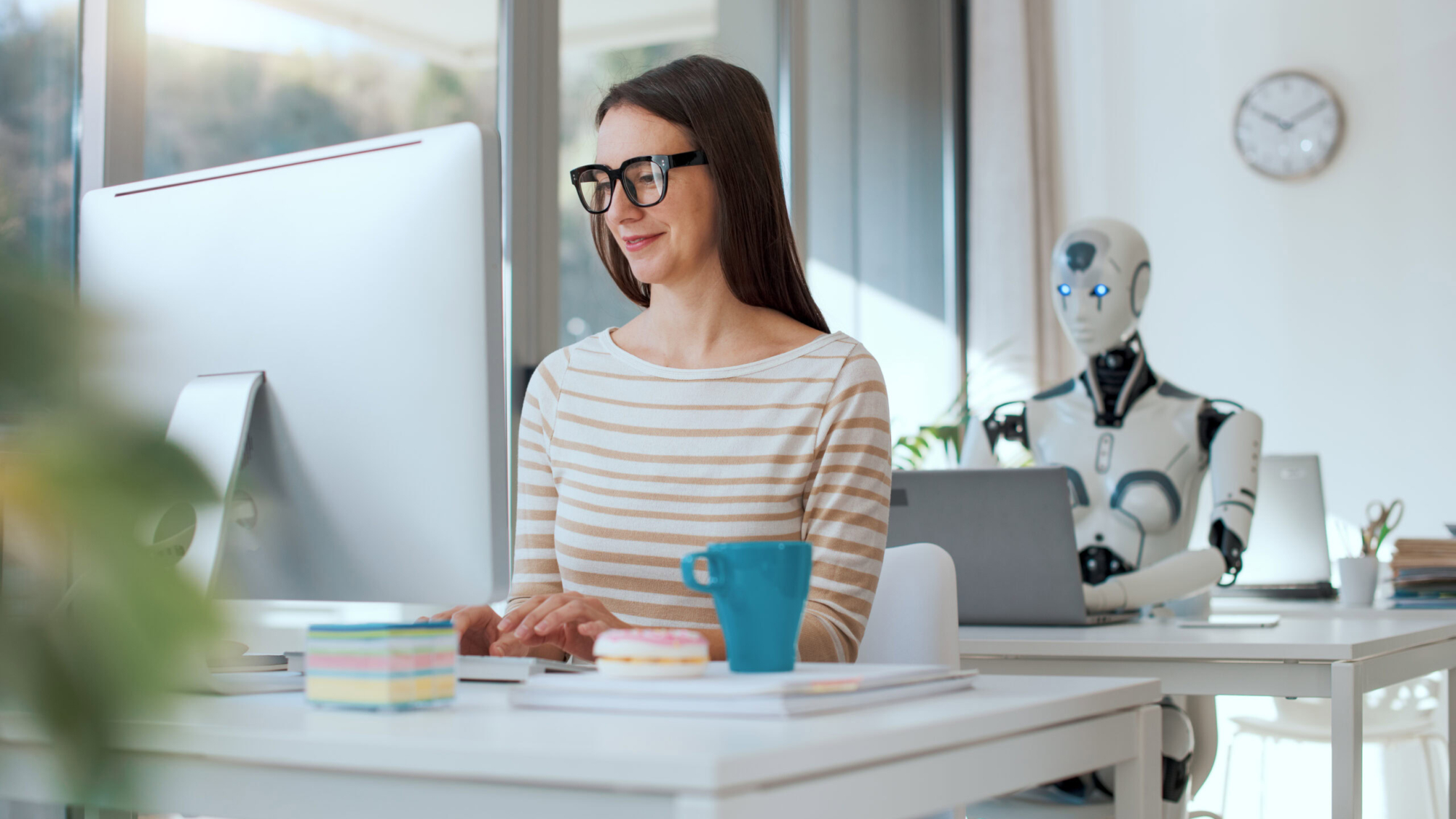 Smiling woman and AI robot sitting at the desk in the office, they are working with computers