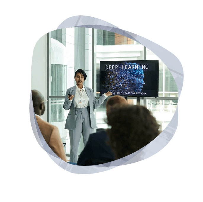 An AI trainer from the Mint Group standing in front of a group, wearing a grey suit, having a workshop on Deep Learning using a monitor.