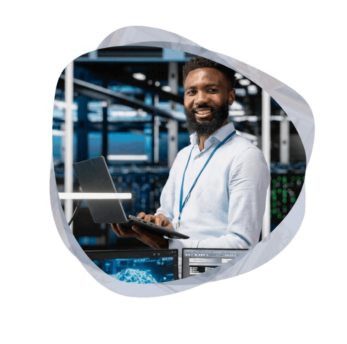 A smiling IT professional in a server room holds a laptop.