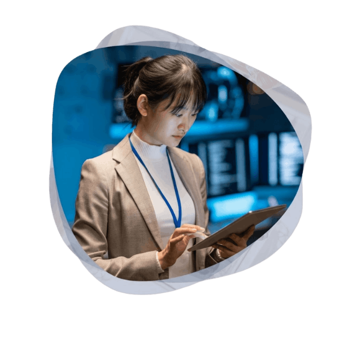 A professional woman wearing a blazer and lanyard reviews information on a tablet in a modern control room with blurred blue digital screens behind her.