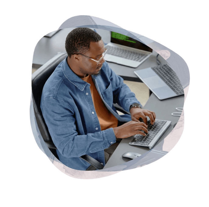 A developer in a blue shirt and glasses types on a keyboard at a multi-monitor workstation while building bespoke customer portal software.