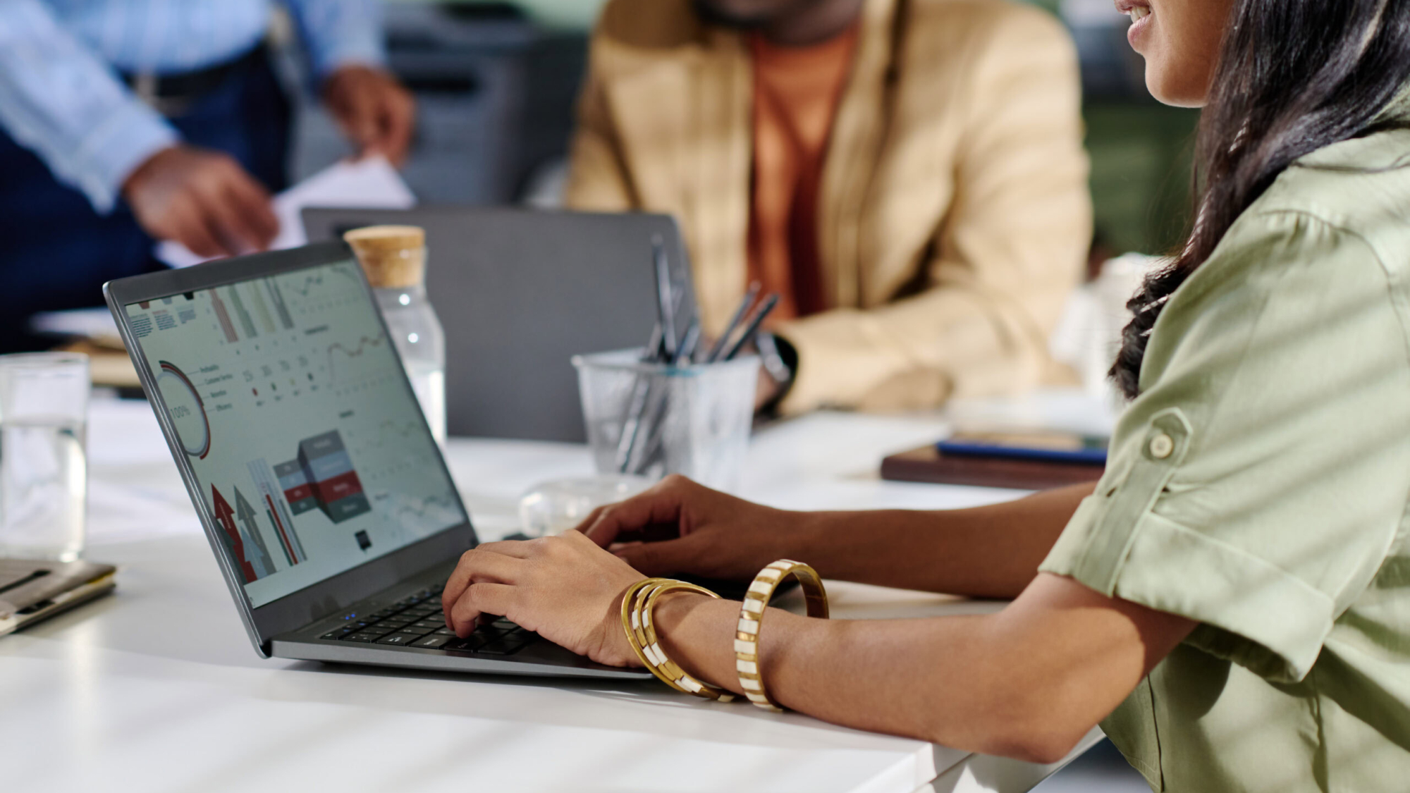Man and woman collaborating in office analyzing business data on laptop screen with charts and graphs surrounding them. Visible documents and coffee cup on table enhancing professional atmosphere