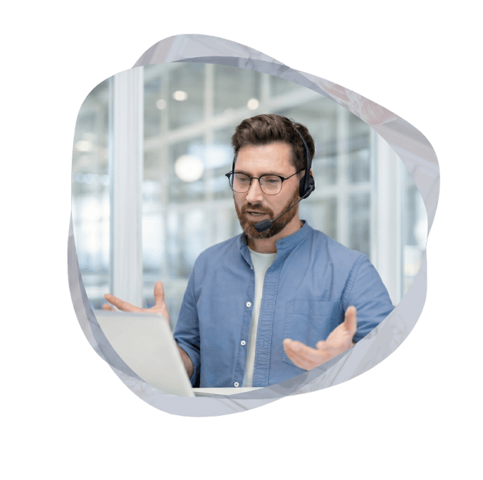 A male customer service agent wearing glasses and a headset, gesturing with his hands while speaking and looking at a laptop.