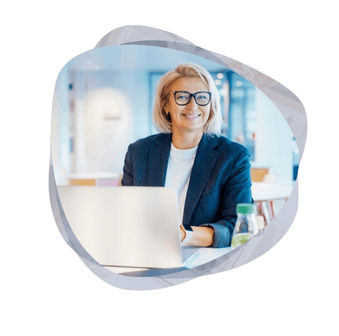 A smiling businesswoman wearing glasses and a dark blazer sits at a desk in a bright, modern office, working on a silver laptop.