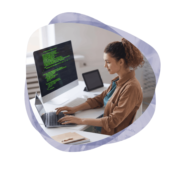 A focused woman typing on a laptop with a desktop monitor displaying code behind her, showing her legacy application modernisation project.