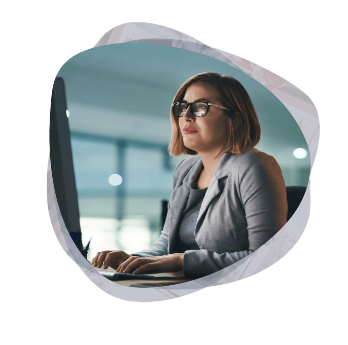 A female professional in a grey blazer working intently at a desktop monitor, establishing governance and compliance rules for a Power Platform Centre of Excellence.