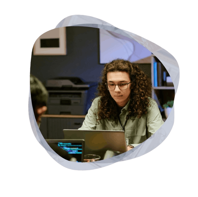 A young man with long curly hair and glasses concentrates on his laptop at a collaborative office desk.