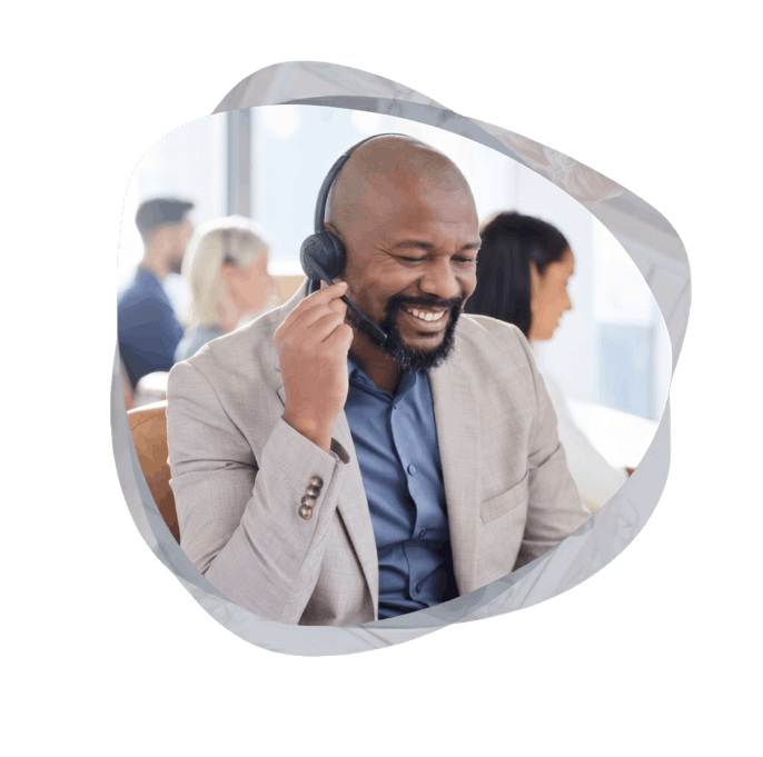 A smiling, smartly dressed male sales professional wearing a headset, looking down happily at his desk in an office environment.