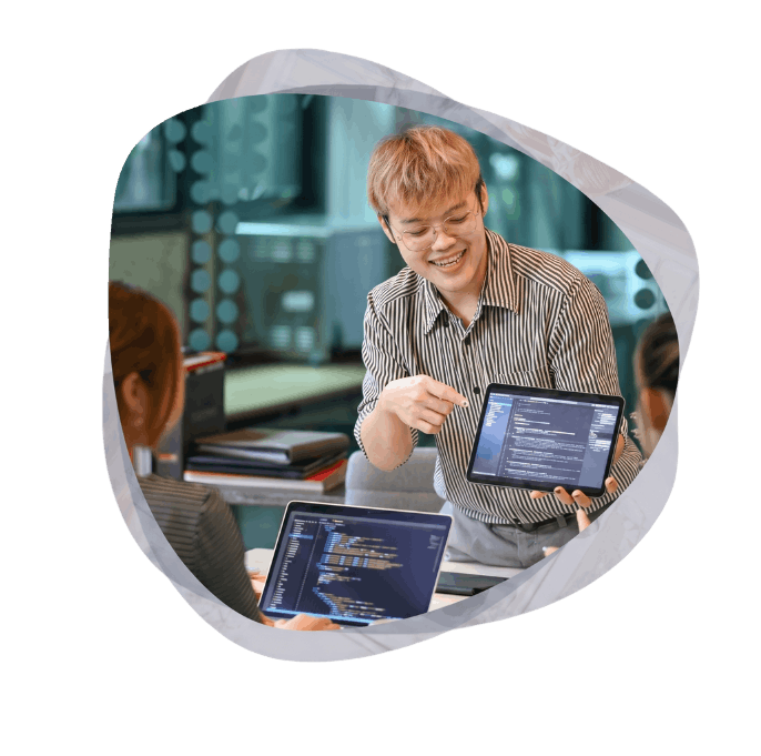 A smiling developer in a striped shirt points to code on a tablet while collaborating with two colleagues, presenting turnkey Power Platform solutions that accelerate deployment.
