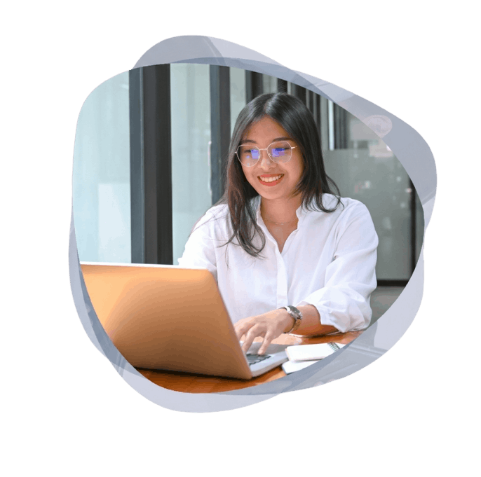 A smiling woman with glasses and a white shirt types on a silver laptop at a wooden desk while performing website platform modernisation and migration.