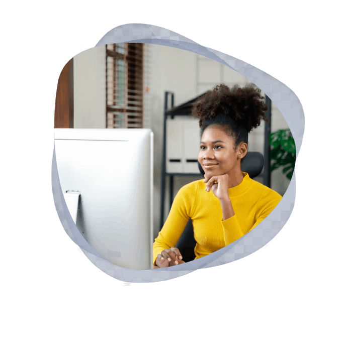 A smiling woman in a yellow turtleneck sits at a desk, resting her chin on her hand as she looks at a desktop computer monitor to learn what is a client portal.