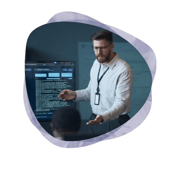 A bearded professional man with glasses and a lanyard points to a large screen displaying computer code while presenting a cloud project to a client.
