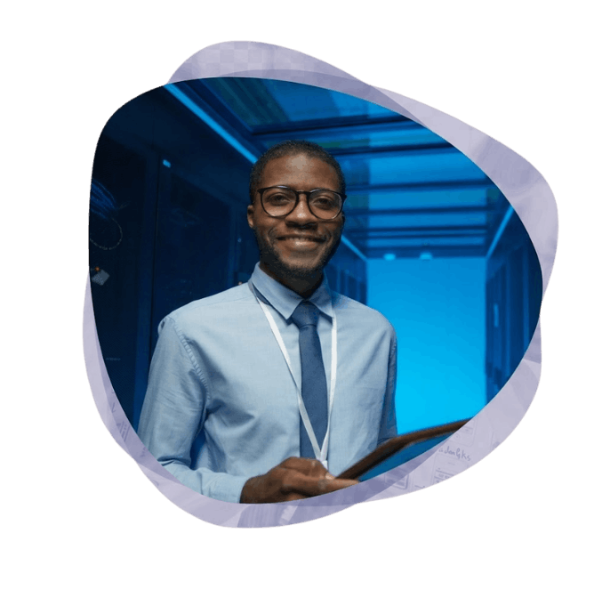 A smiling man with glasses, a light blue shirt, and a tie holds a tablet while standing in a data center aisle illuminated by bright blue light, framed within a soft purple amoeba-shaped border.