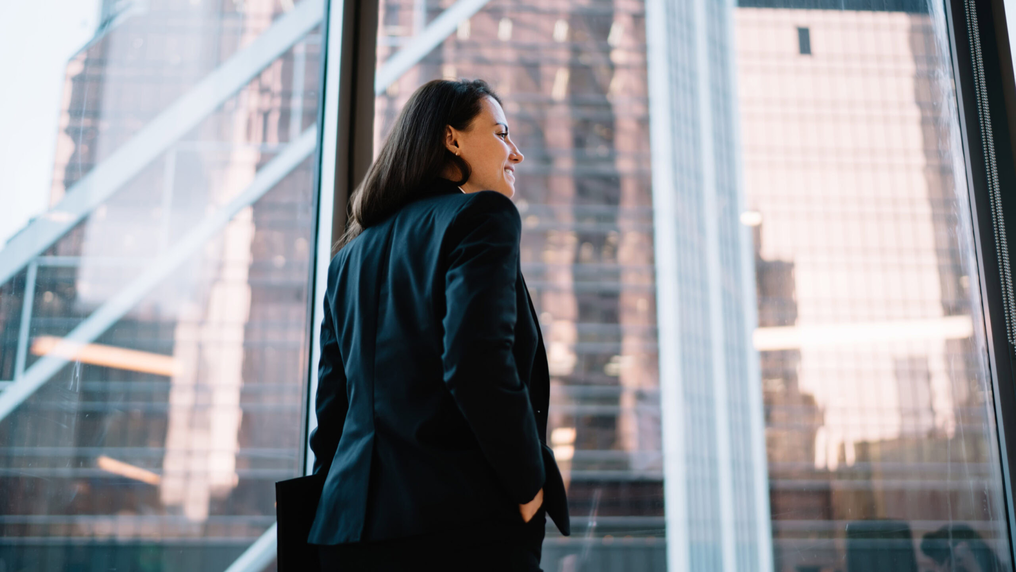 Low angle side view of young female in black formal outfit with hand in pocket standing against panoramic window and enjoying downtown view